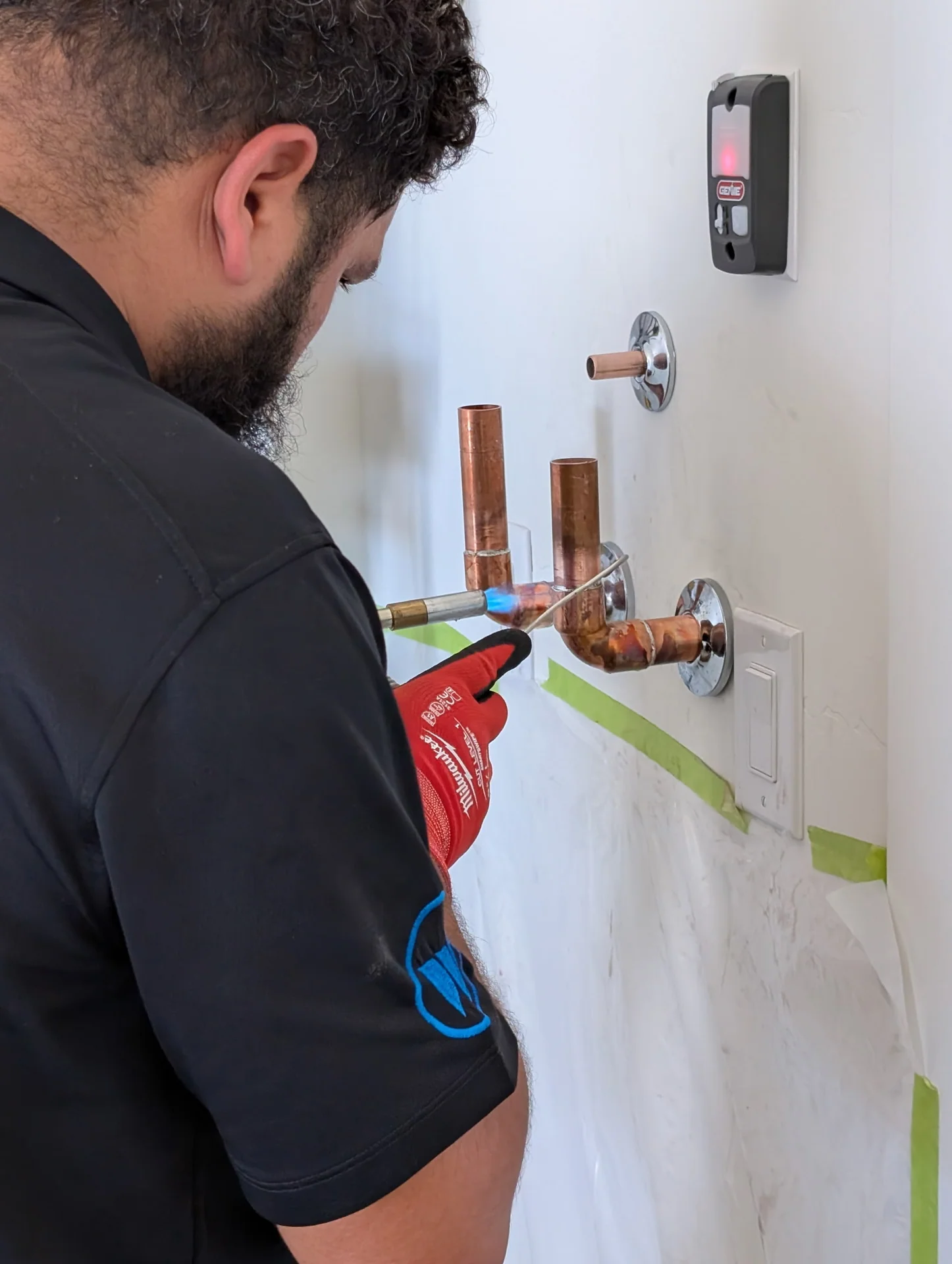 Ancla plumber soldering copper supply lines for a shower valve, taped-off drywall, red gloves, propane torch in hand