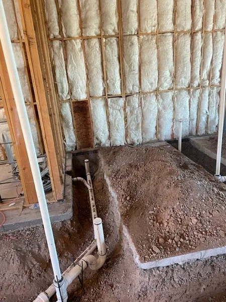 PVC drain rough-ins in a sandy trench next to spray-foam insulated walls