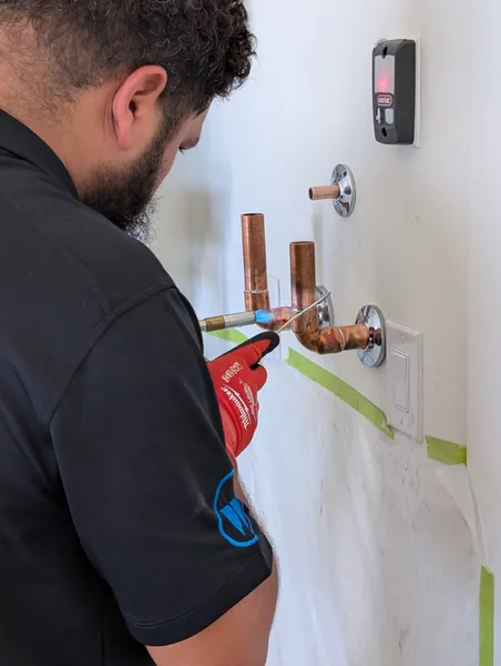 Ancla plumber soldering copper supply lines for a shower valve, taped-off drywall, red gloves, propane torch in hand